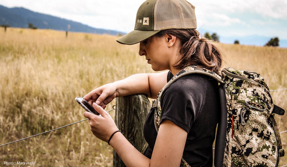 Woman looking at phone while hunting and hiking beside fenceline.