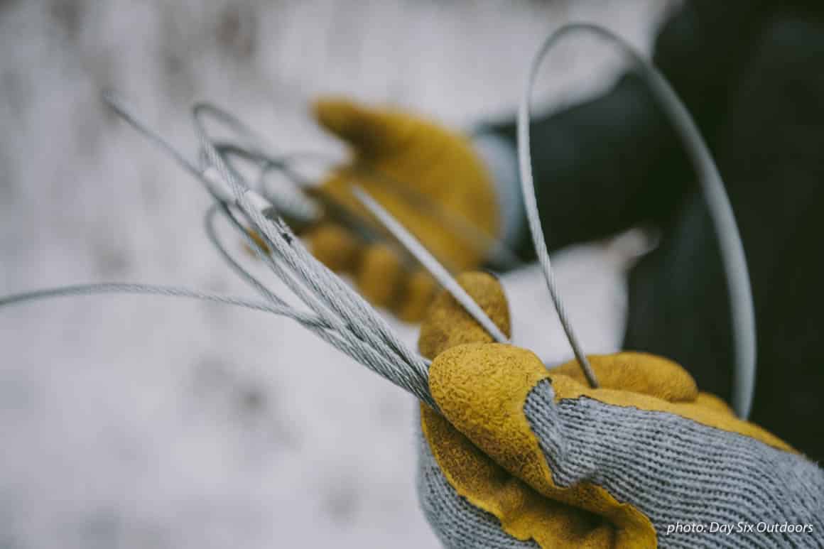 Man setting up snare trap in the winter, wearing gloves in the snow, for animal trapping.