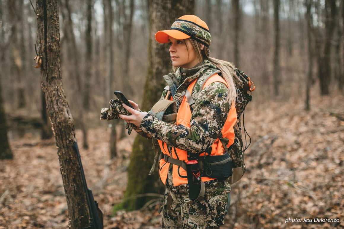 Woman hunting in woods in camouflage with cell phone.