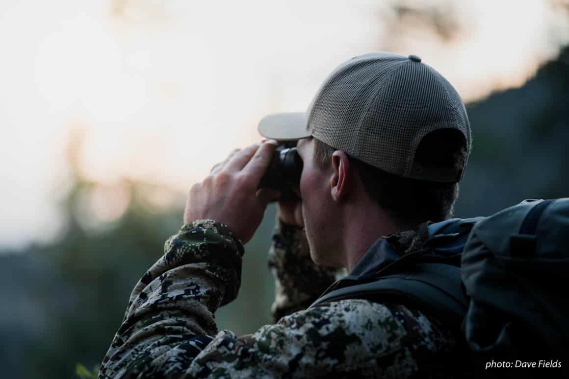Man in camouflage uses binoculars to spot elk while hunting in the Montana backcountry.