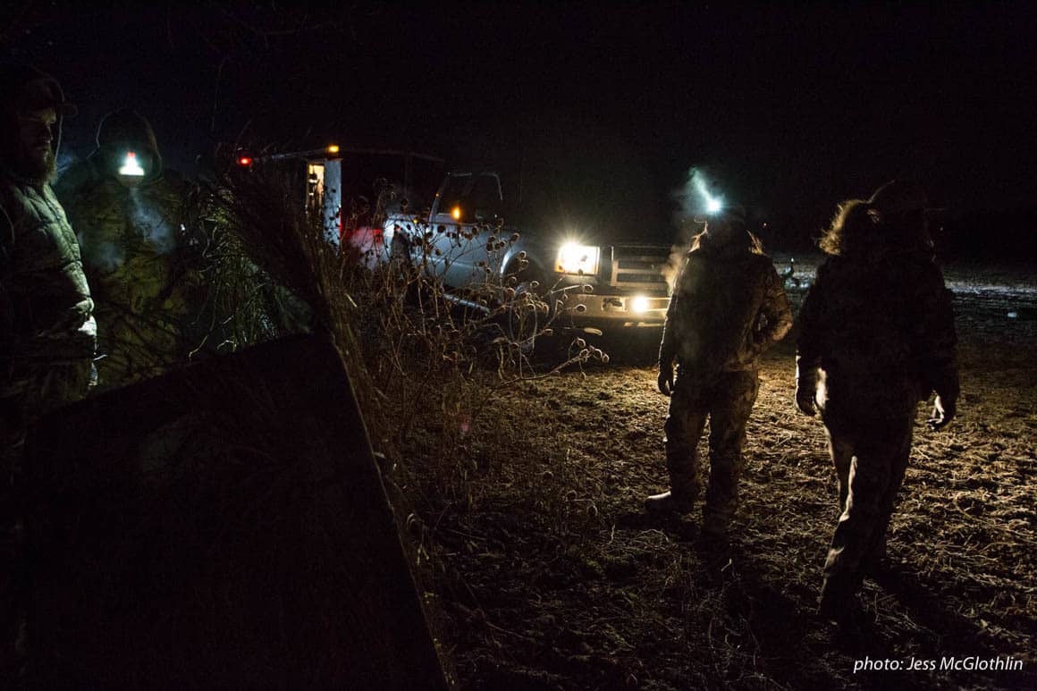 Waterfowl hunters set up decoys and a blind in darkness by the light of truck headlights.