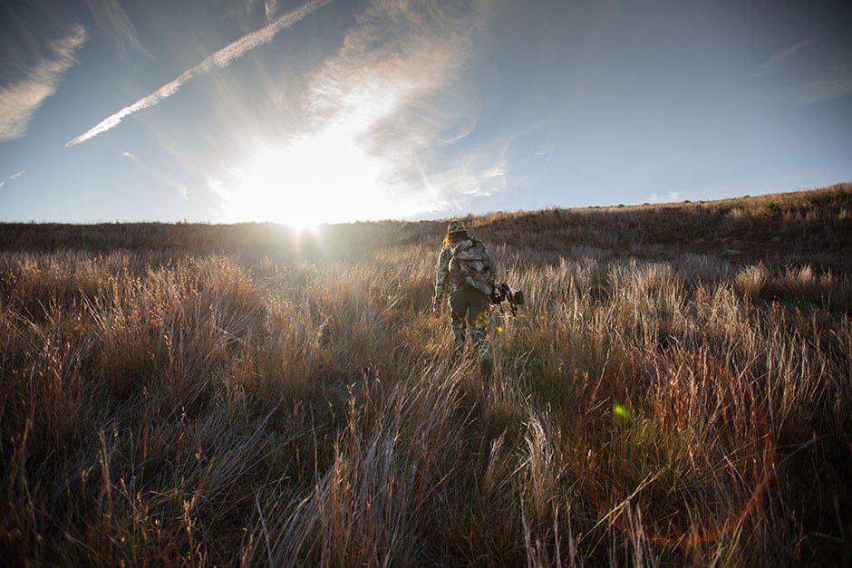 Image of female hunter in camo with a bow walking into the sunset in a field on Western public lands.