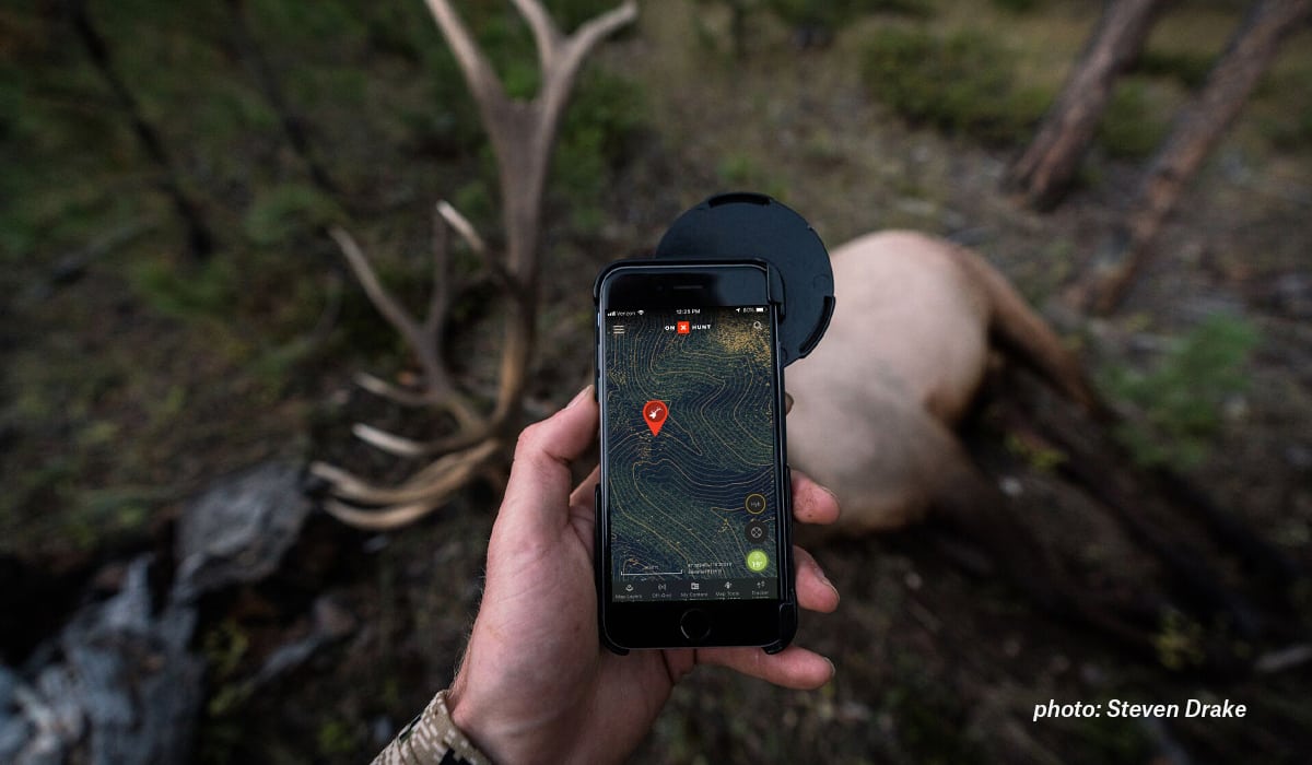 A hunter stands over an elk looking at onX Hunt with an elk Waypoint.