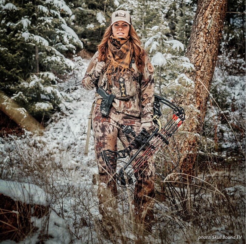 Woman in camouflage hunting in the snowy woods holding a compound bow.