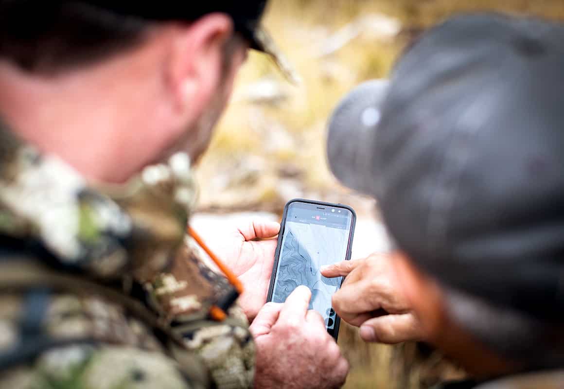 Two men look at a phone displaying the onX Hunt App while scouting in the woods and hunting.