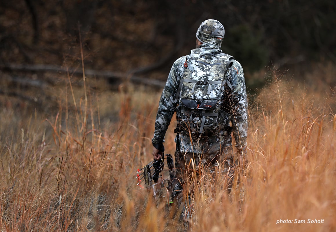 Image of a man in camouflage walking through autumn grass with a bow in hand while hunting.