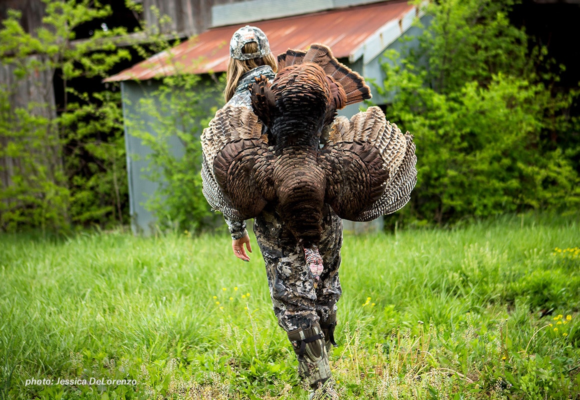 Woman in camo walking with turkey over her shoulder after hunting.