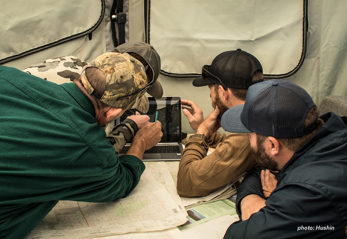 Men gathered in a tent around a computer, e-scouting on onX Hunt Web Map before they go hunting.