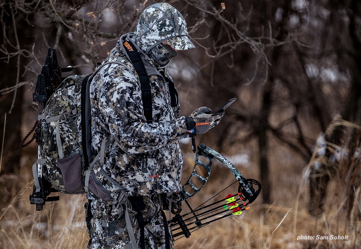 A man in full camouflage and a compound bow checks his onX Hunt App on his mobile phone while hunting.