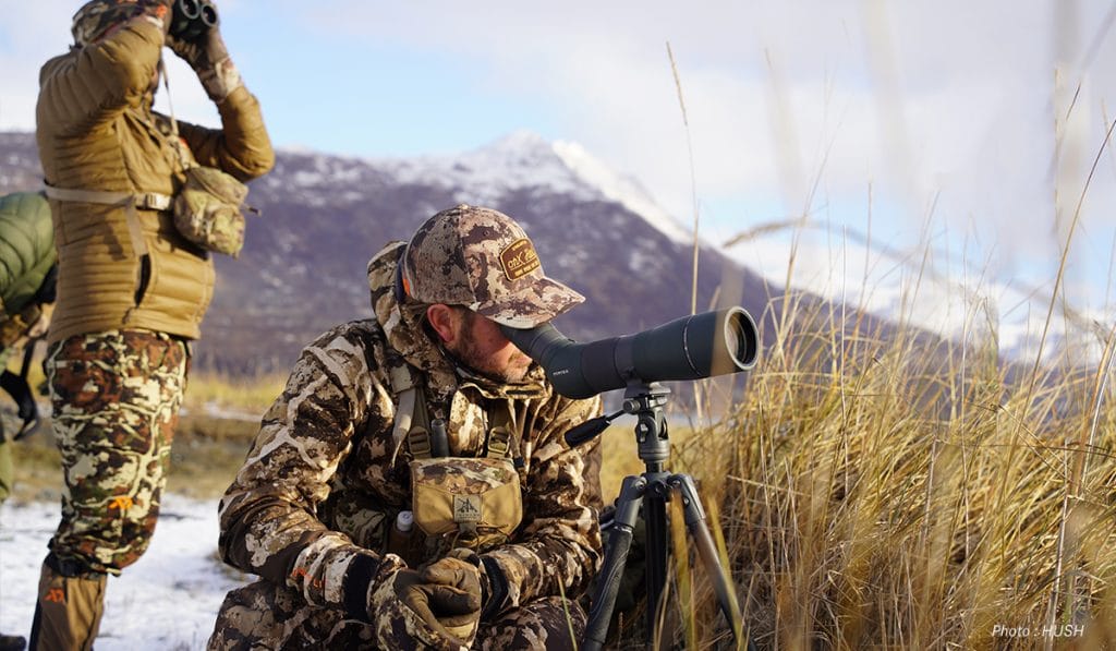One hunter stands and uses his binoculars to glass while another hunter sits and uses a spotting scope. 