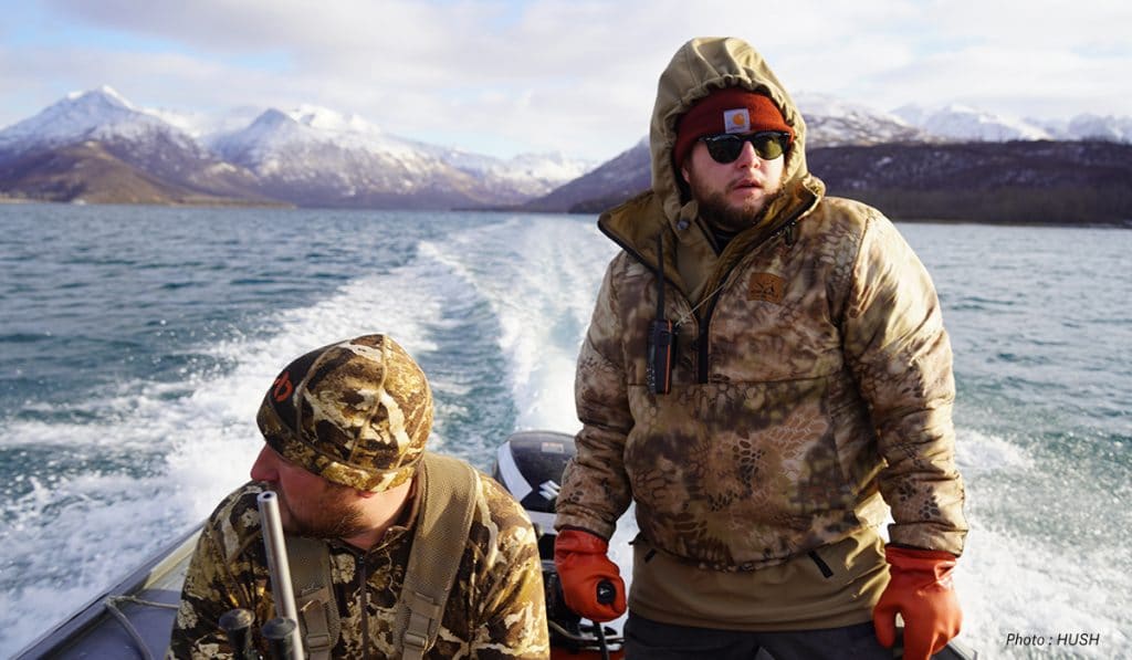 Two men in camo on a boat with snowy mountains in the background. 