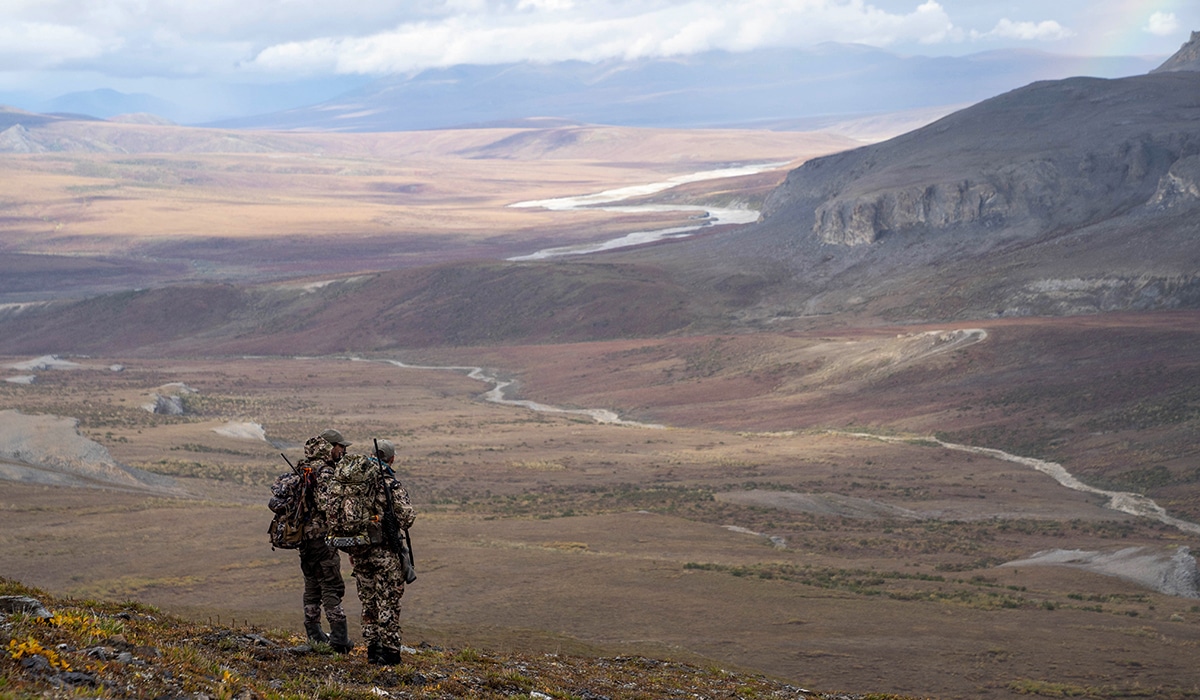 Two hunters standing on a mountain in Alaska overlooking a drainage.