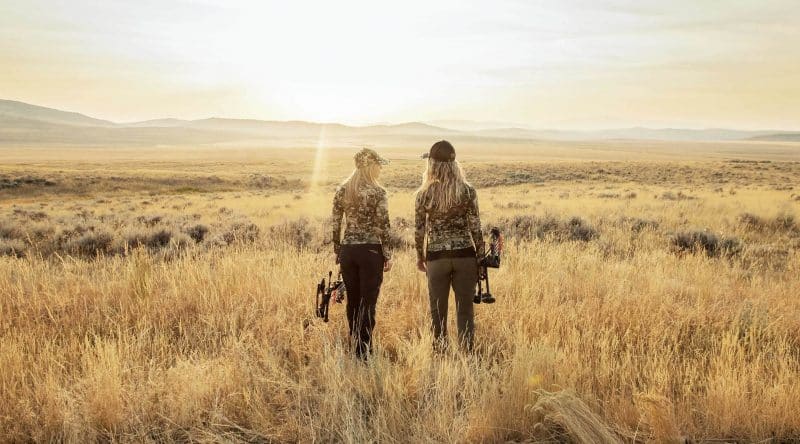 Two female hunters stand in the prairie.