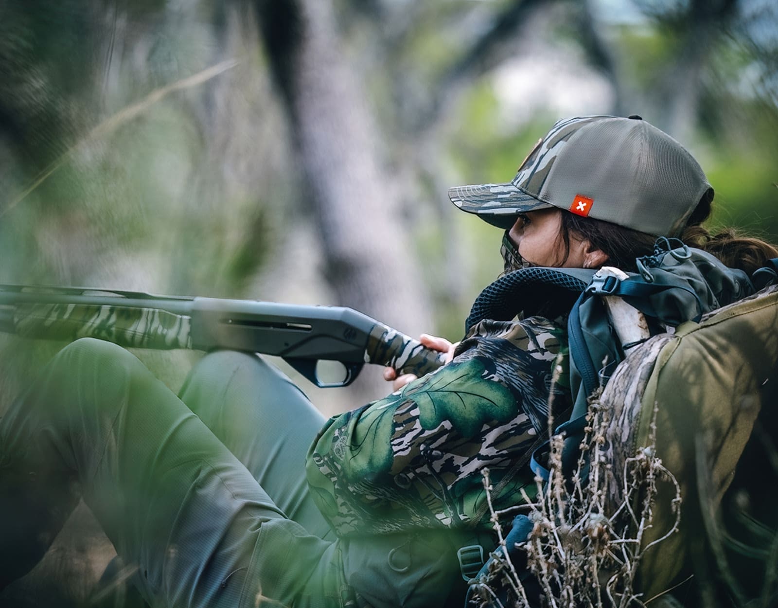 A female turkey hunter leans against a tree holding a shotgun.