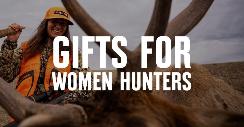 A female hunter in blaze orange smiles while looking at a bull elk.