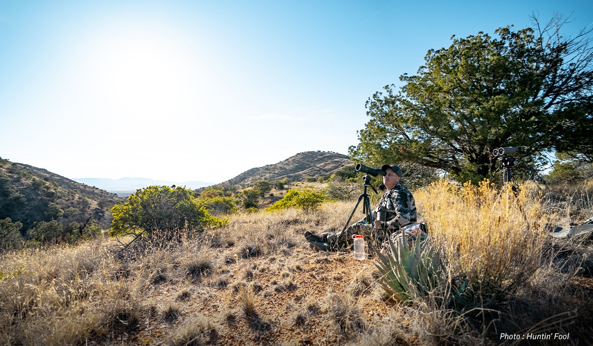 A hunter sits on the ground and uses a spotting scope to glass for game on the next mountain ridge.