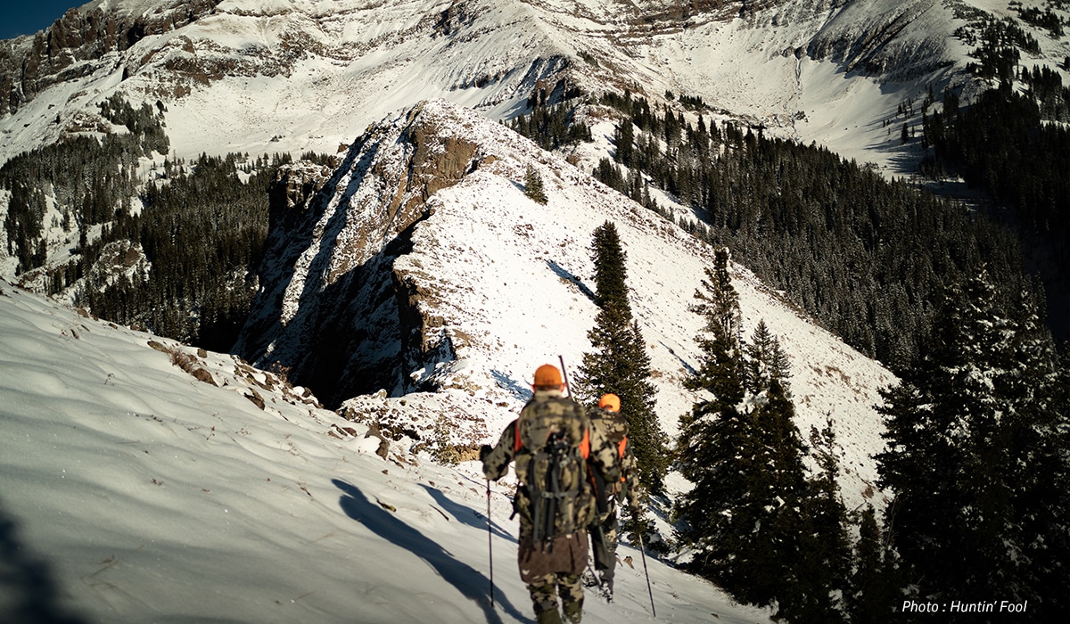 Two hunters use trekking poles to navigate a snowy mountain descent. 