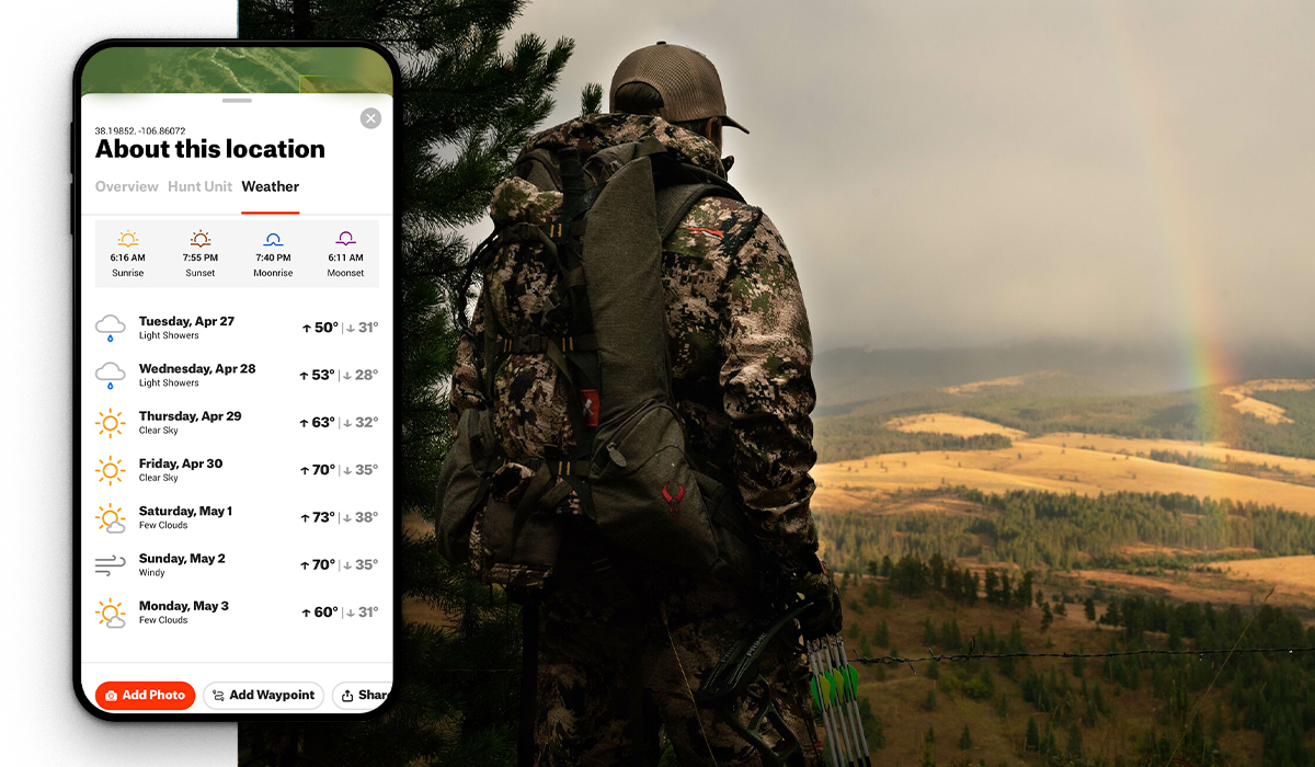 A bowhunter stands on a hillside looking out over a meadow with a rainbow overhead. A screenshot of the onX Hunt App shows the weather user interface. 