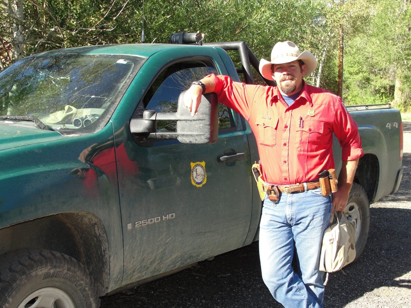A game warden in a red shirt and cowboy hat poses for a photo in front of his green work truck.