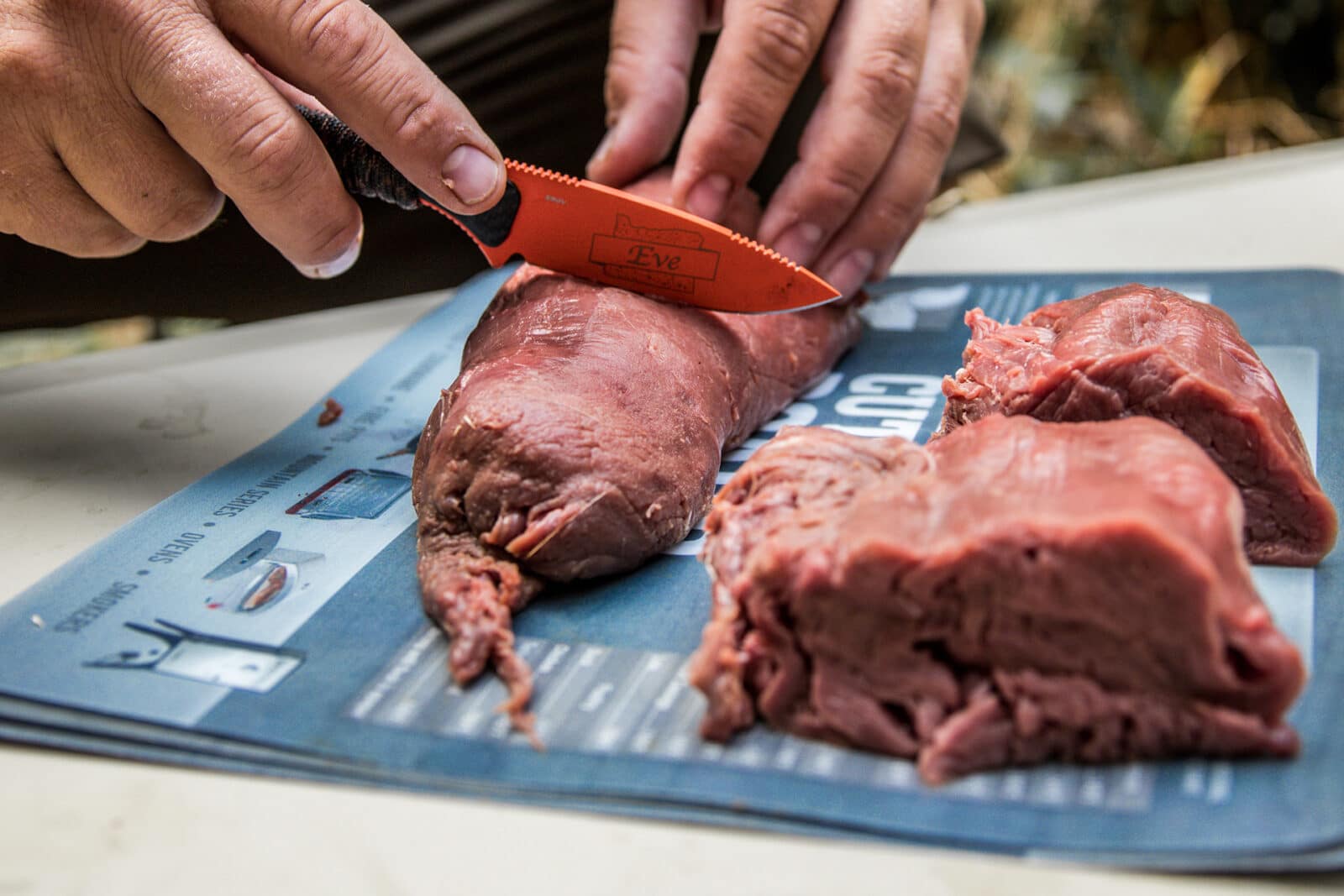 Hands using an orange knife to cut into wild game meat.