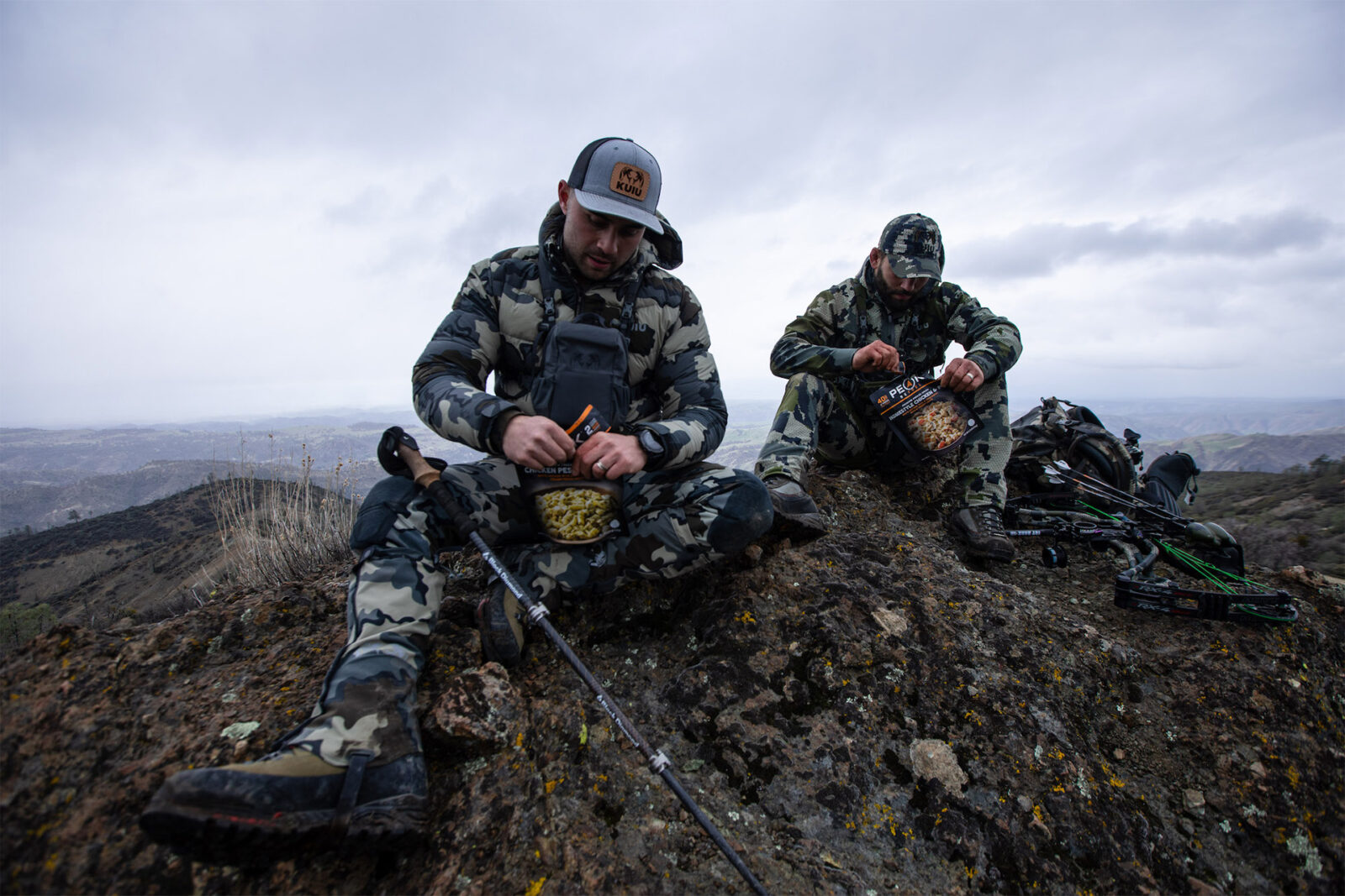Two hunters sit on top of a mountain eating Peak Refuel. 