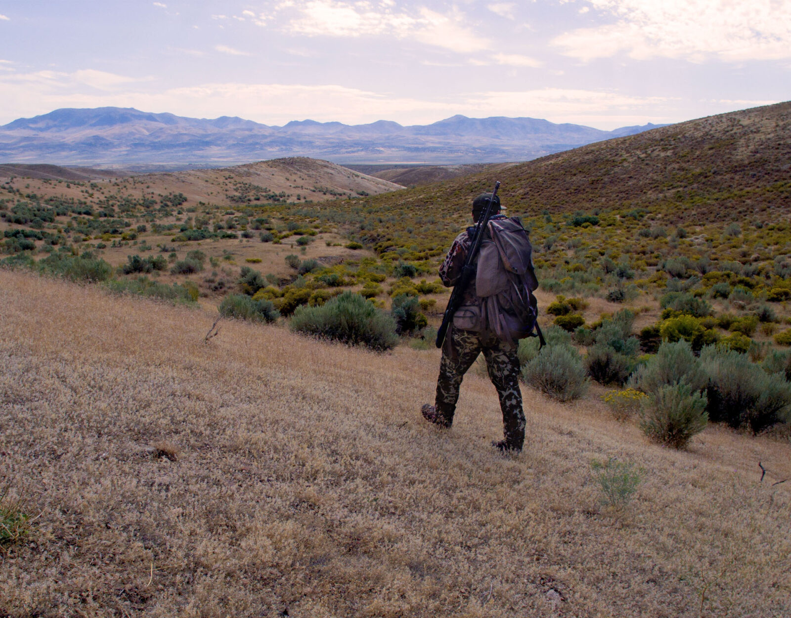 A hunter with a pack walks in a high-desert mountainous landscape. 