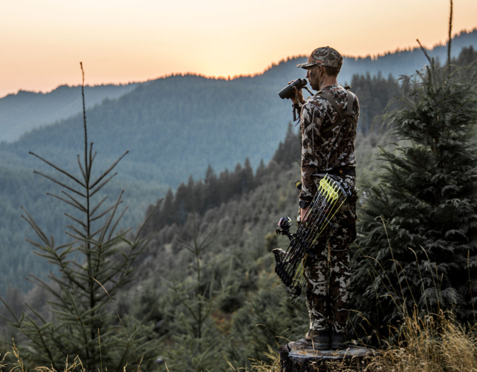 A backcountry hunter holds his bow in one hand and binoculars in the other, facing out into a vast swath of mountainous timber. 