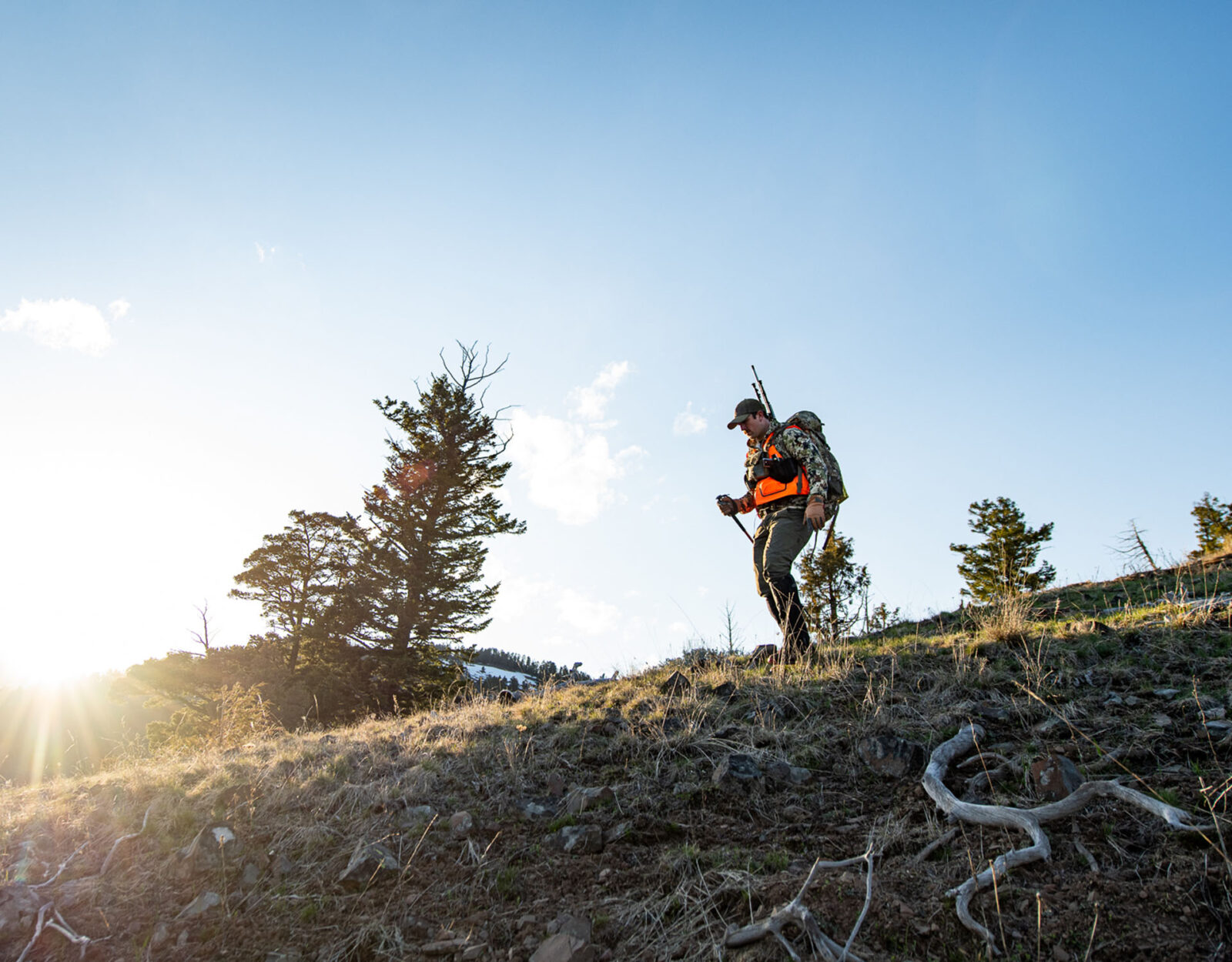 A hunter in blaze orange and camo hikes down the mountain. He is silhouetted by blue skies. 