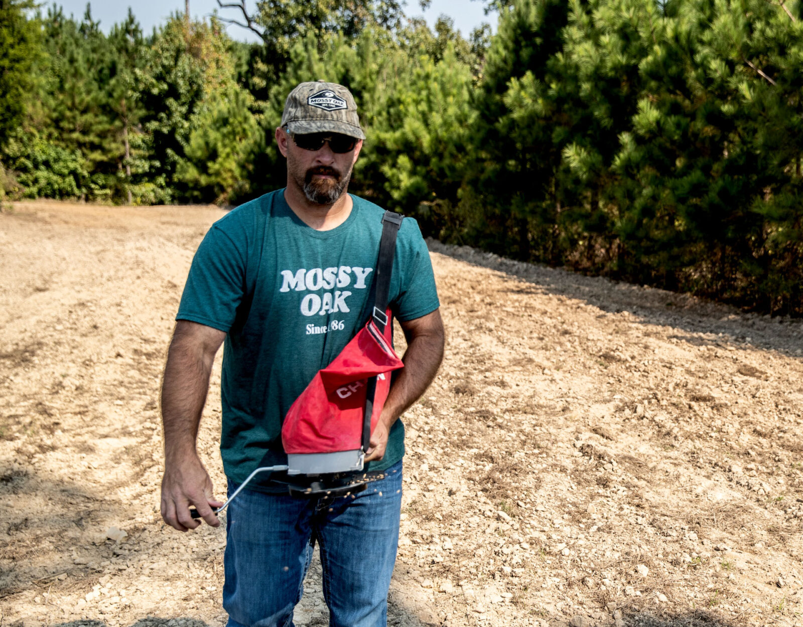 A man in a Mossy Oak tshirt uses a handheld seed spreader. 