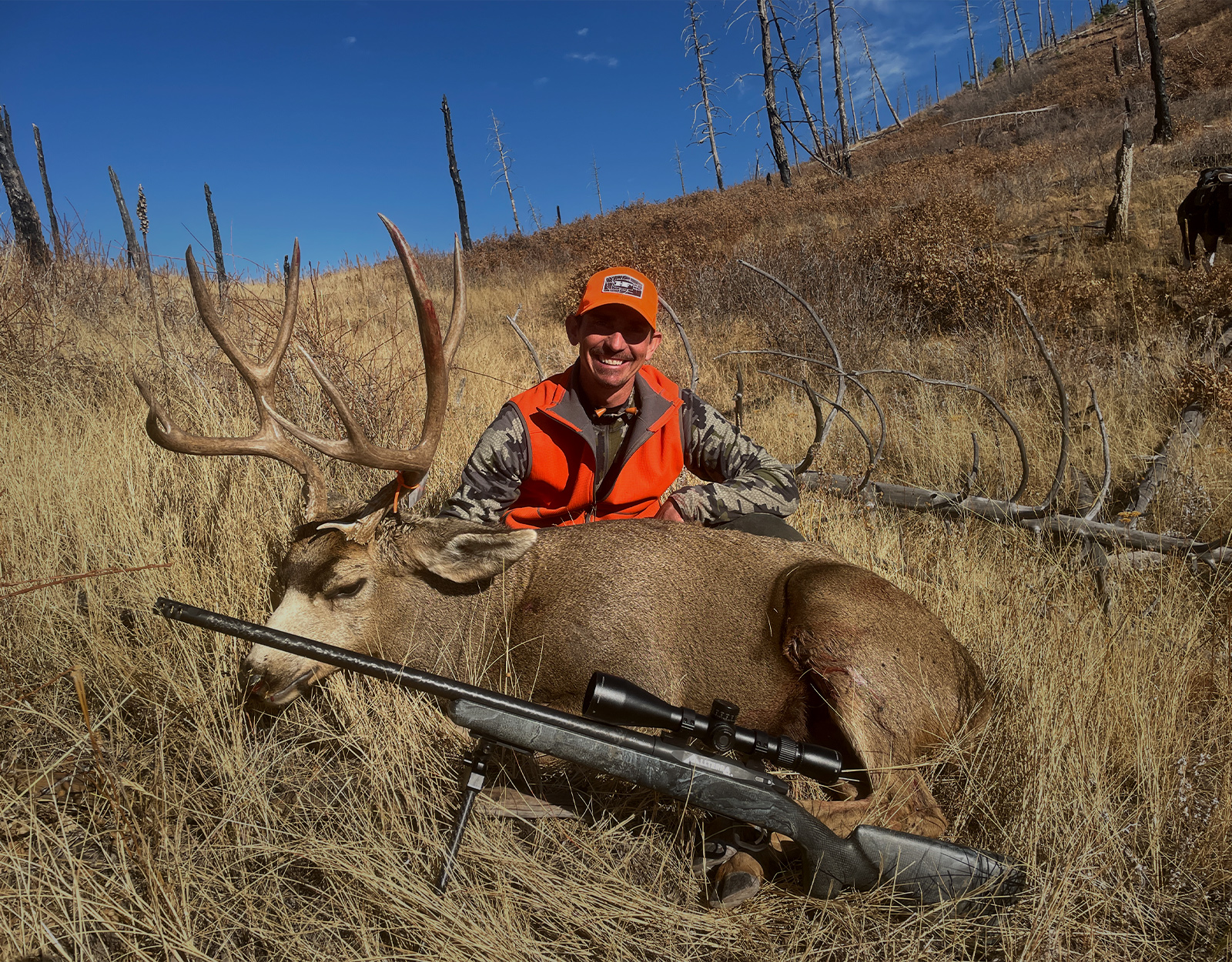 A hunter in blaze orange poses with a mule deer buck he harvested with his rifle. 