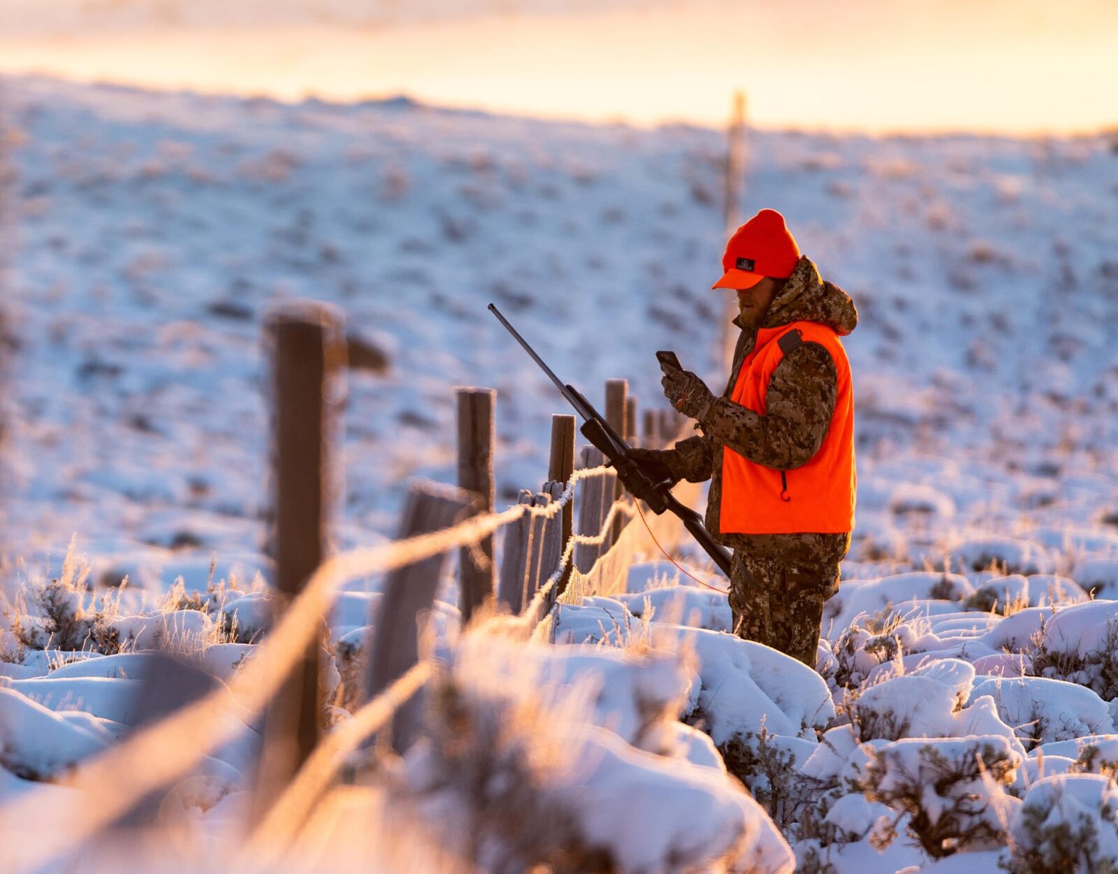 a hunter wearing hunters orange and carrying a rifle while looking at their onX Hunt app on their phone