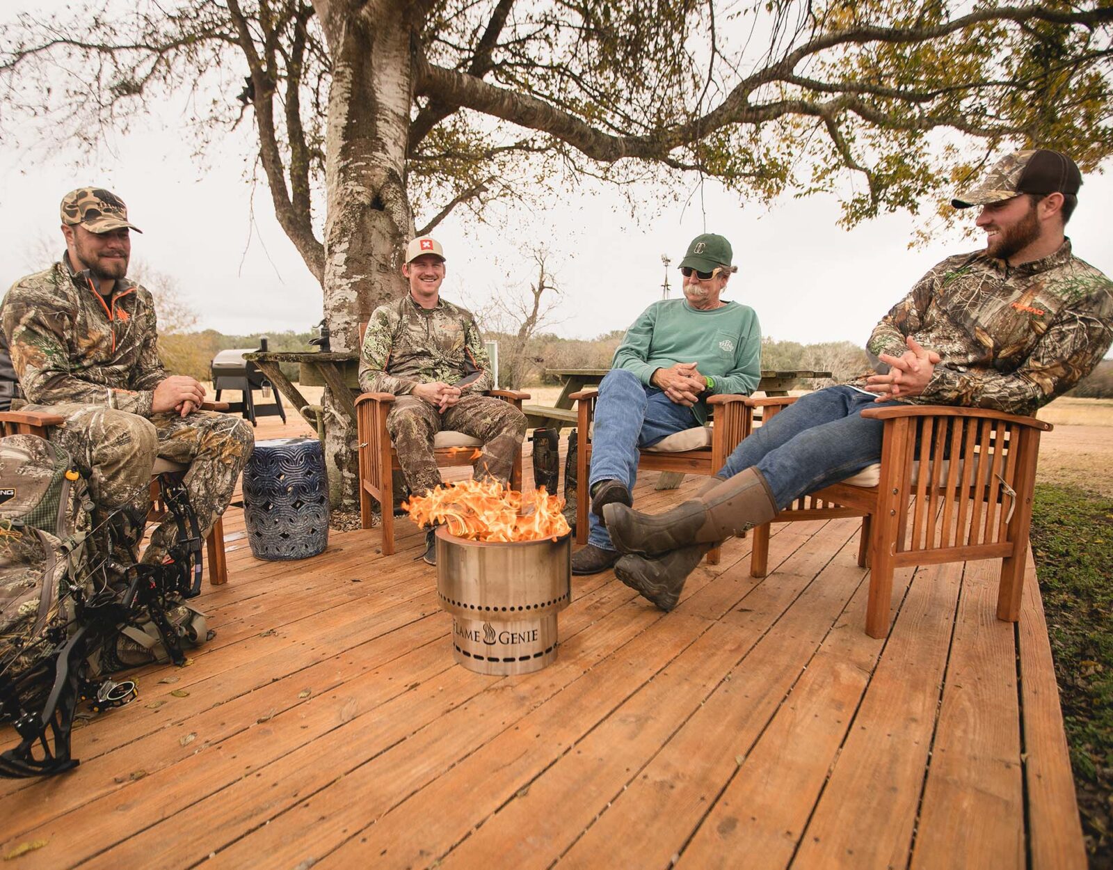 5 men in camo and hunting gear relaxing on a deck with a fire pit.
