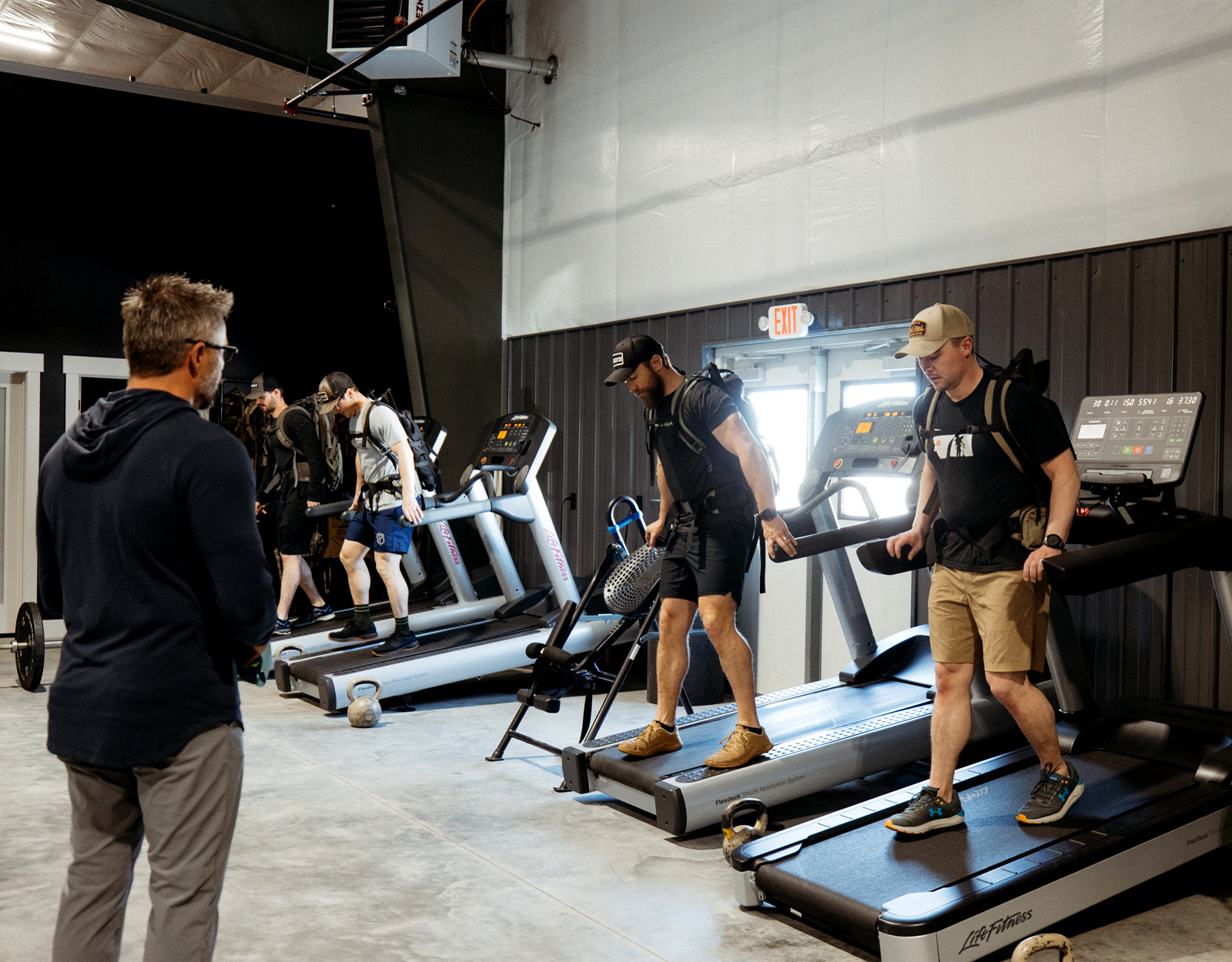 Four men walk backward on inclined treadmills wearing ruck sacks. 