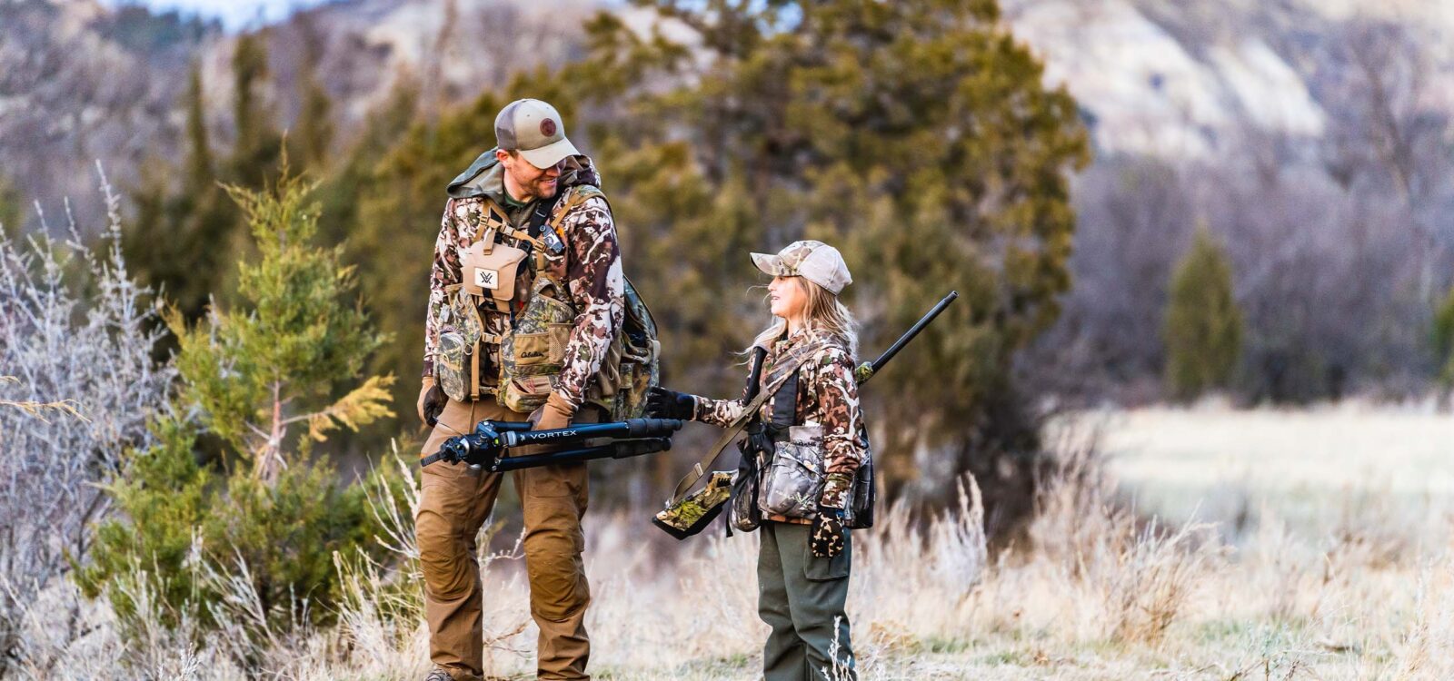 A dad and daughter in camo walk side by side with foliage in the background.