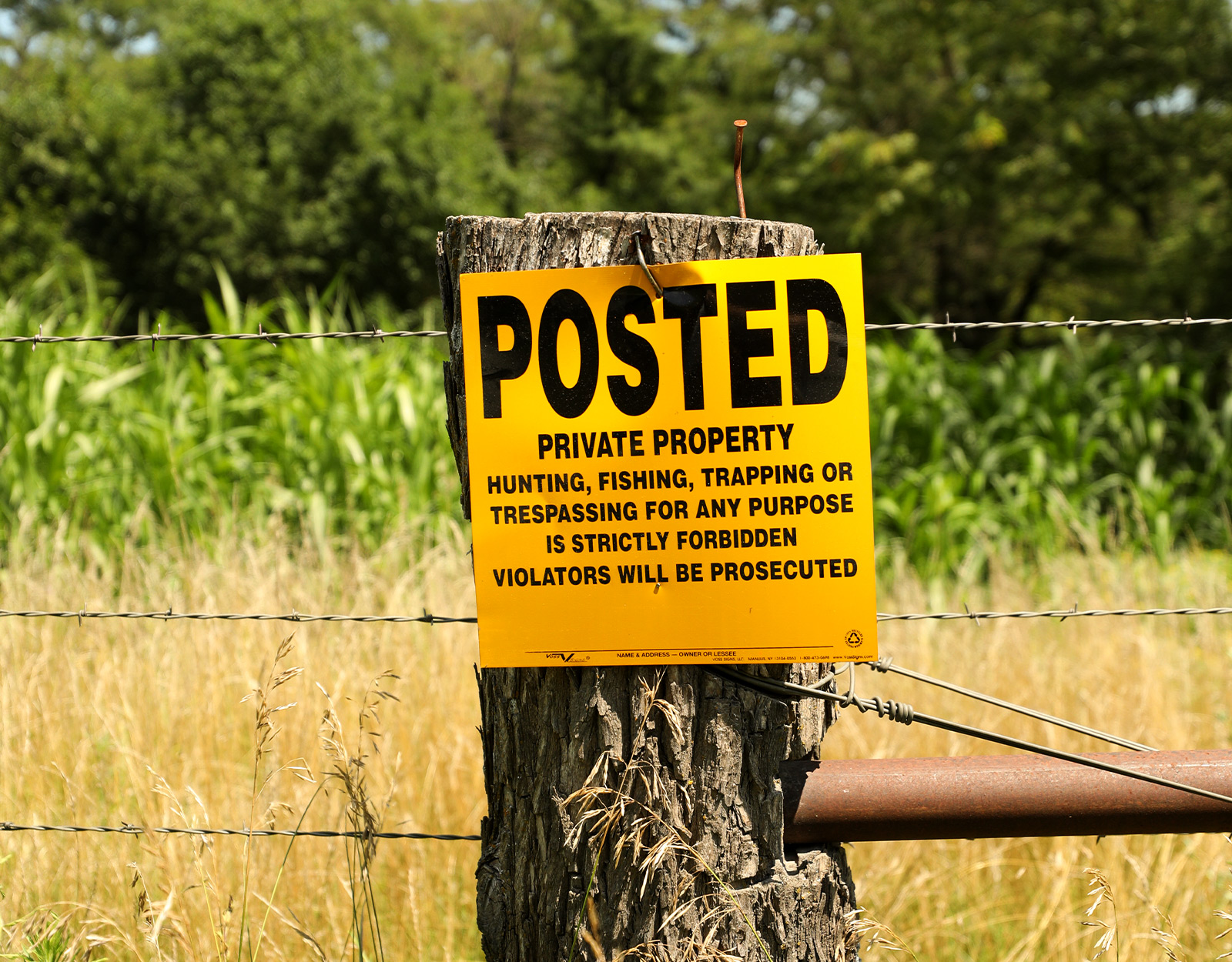 A yellow private property sign is posted to a barbed wire fence. The sign reads: "Posted. Private Property. Hunting, fishing, trapping, or trespassing for any purpose is strictly forbidden. Violators will be prosecuted."