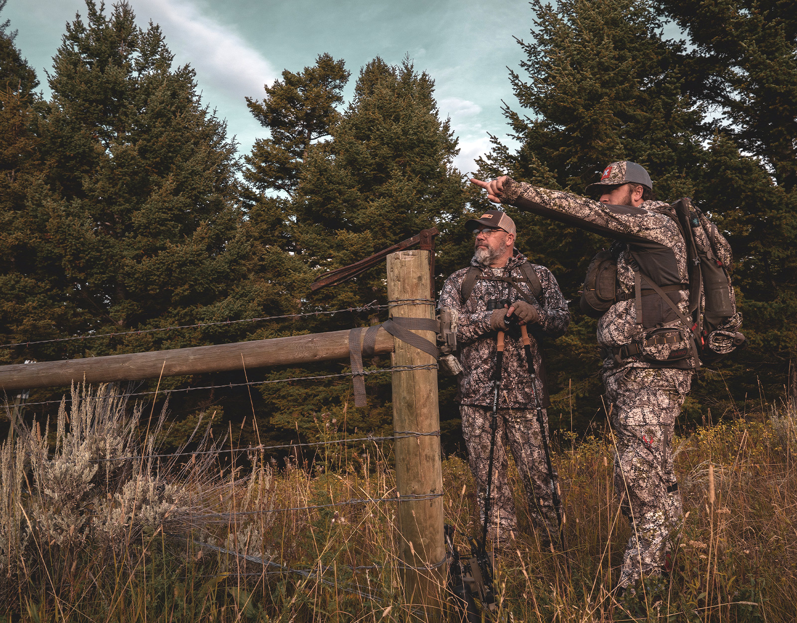 Two male hunters stand by a fenceline. One is pointing at something in the distance. 