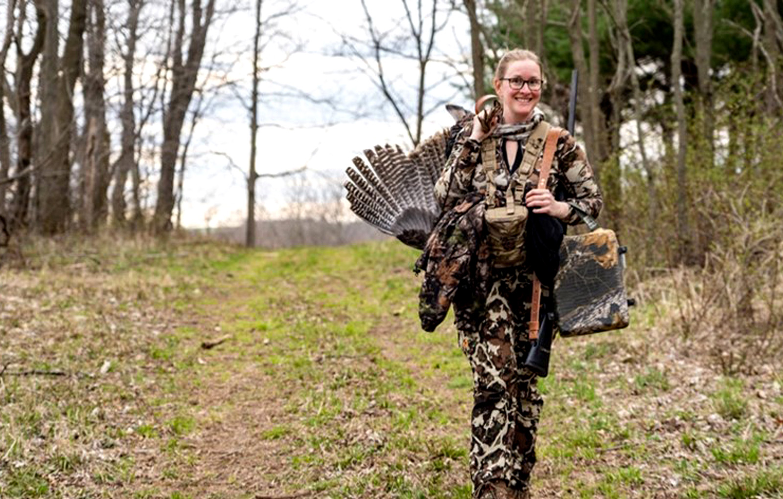 Woman in camo walking with the turkey she harvested over her shoulder.