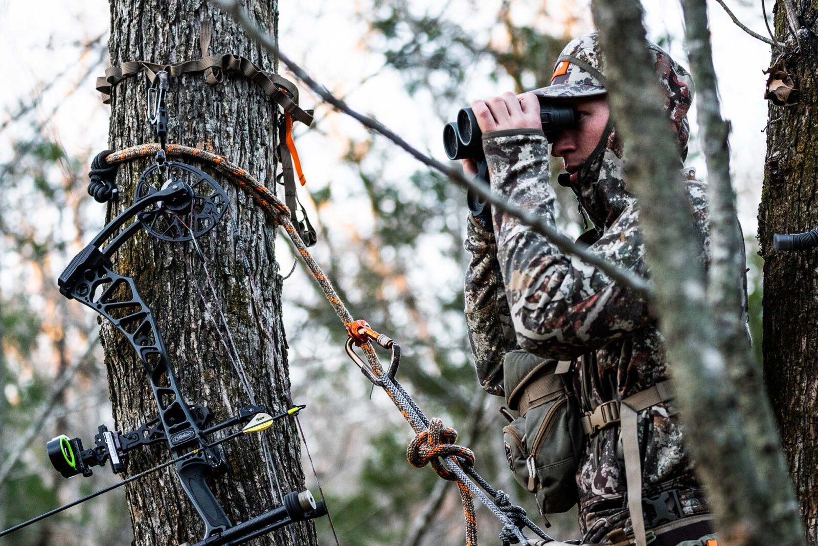 a hunter with binoculars in a treestand with ropes and pulleys holding his gear 