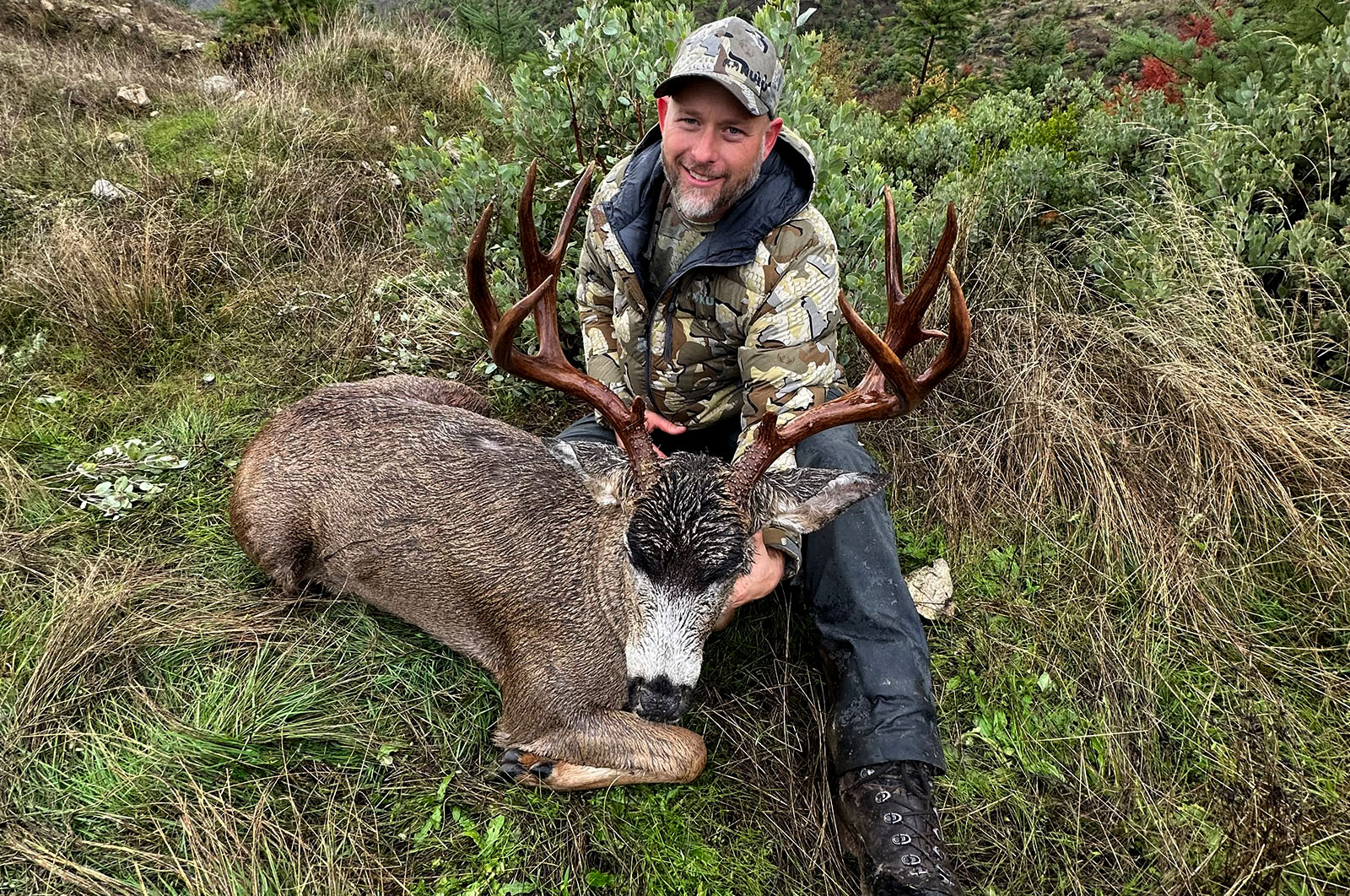 A hunter in camo with the blacktail buck he harvested in a field. 