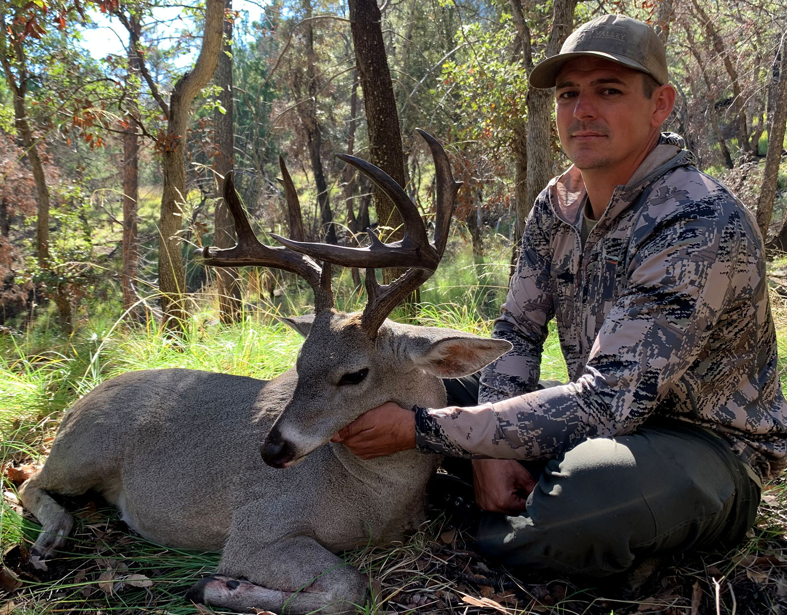 A male hunter in camo with the Coues deer he harvested in a forested area.