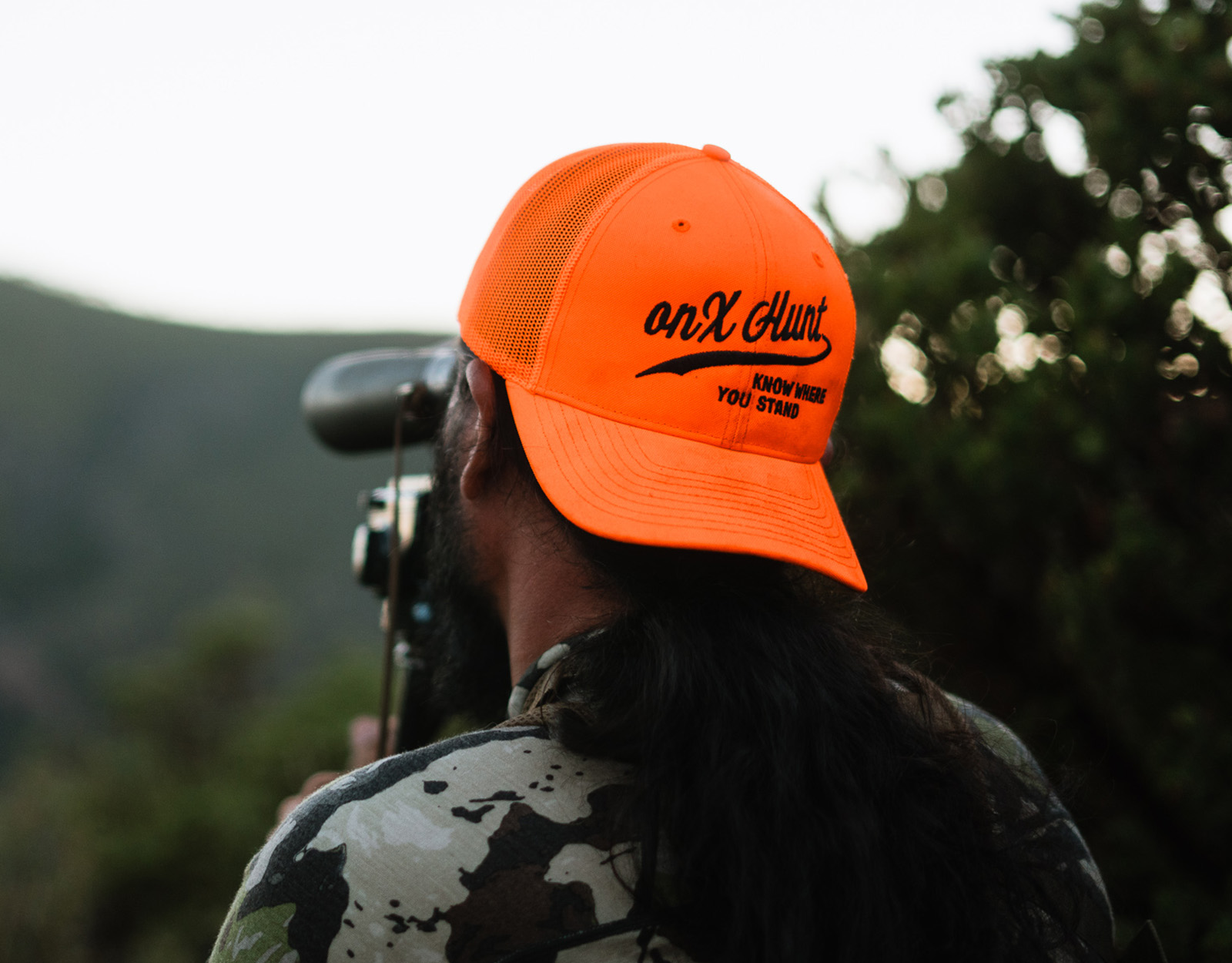 A hunter looking through binoculars with an onX Hunt hat on 

