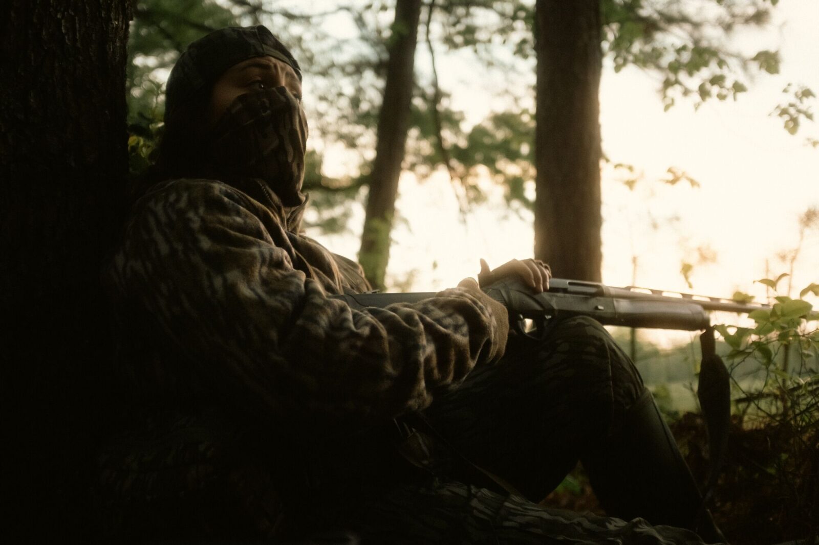 A woman sits in full camo next to a tree.