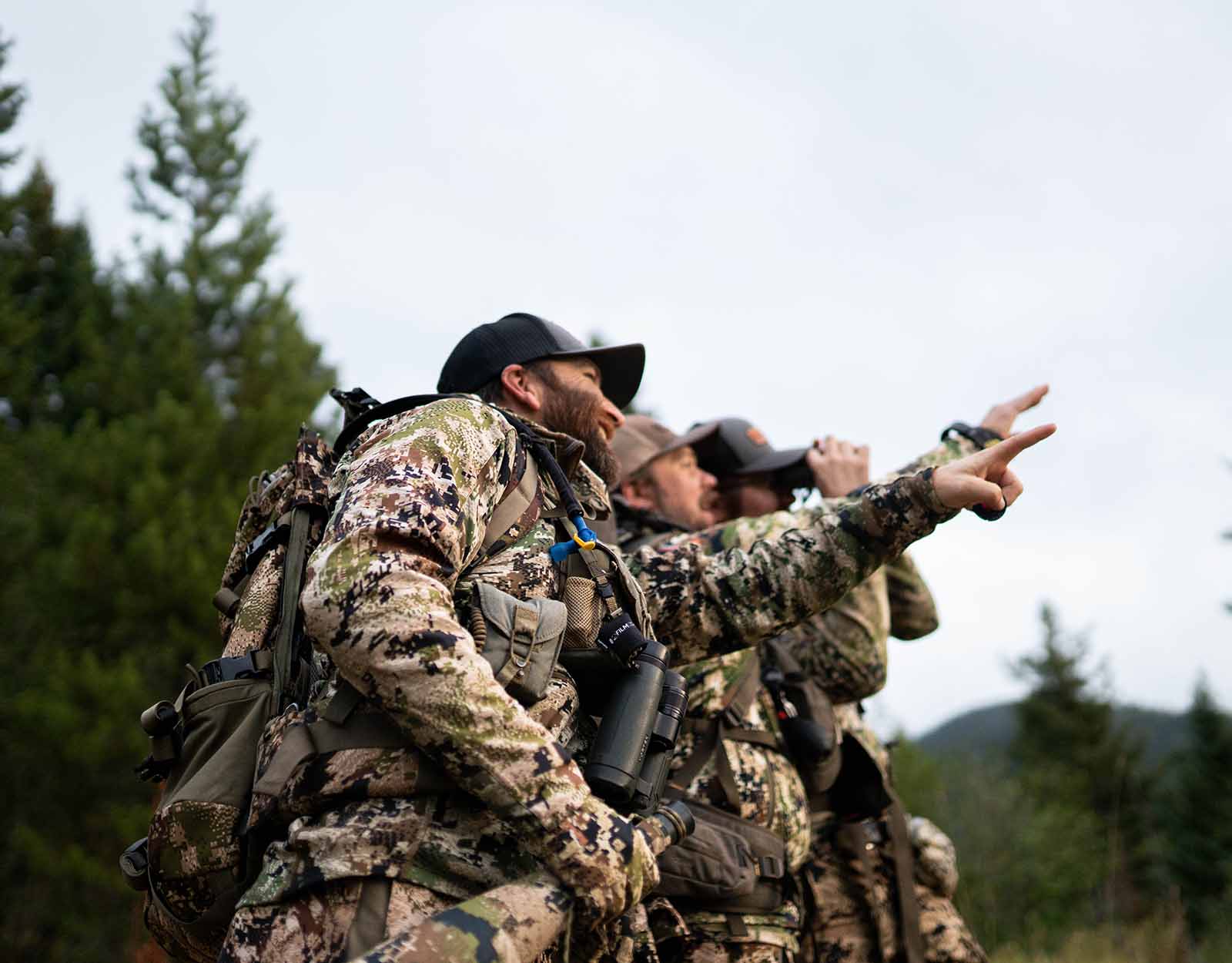 Three men in camo stand in a line, two of them point at something in the distance. 