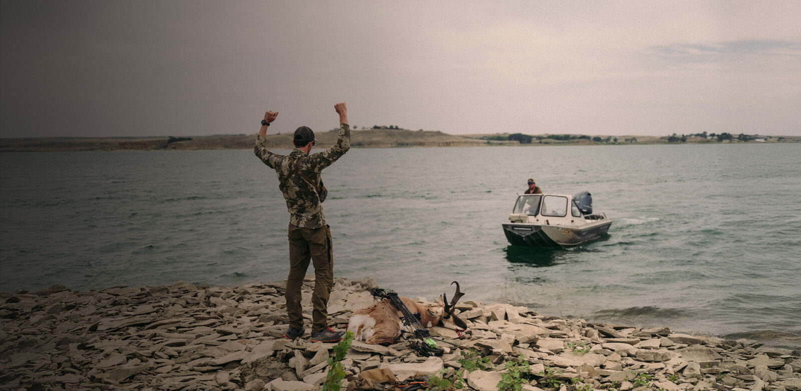 A person stands at the edge of water with a boat approaching them, and celebrates their antelope harvest.