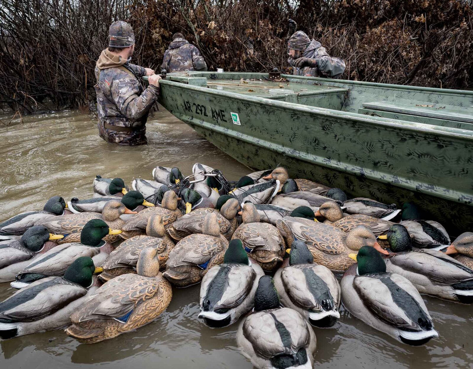 Three hunters stand waist deep in water with a boat and duck decoys. 