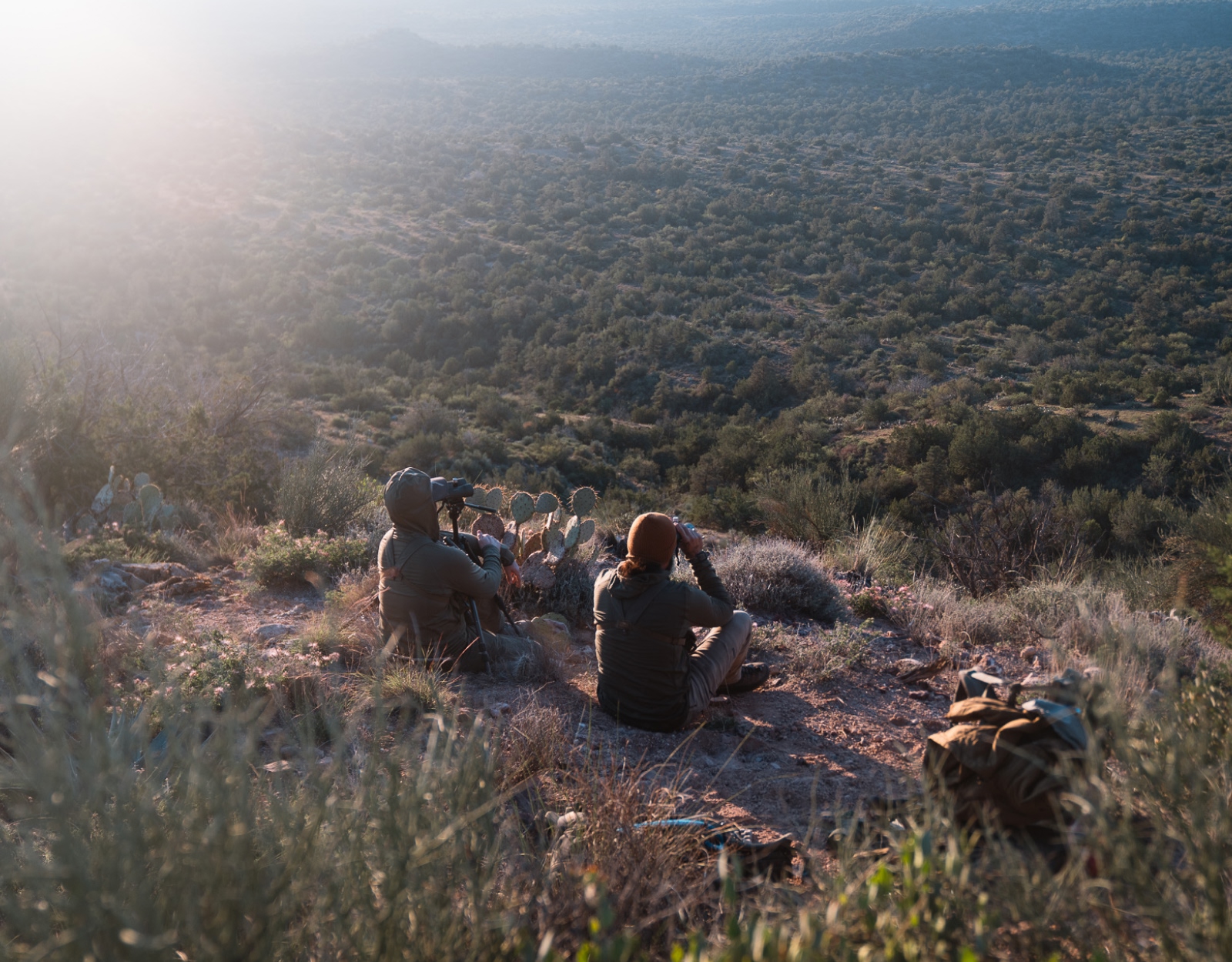 Two men sit in the high desert using binoculars to explore shrubland. 