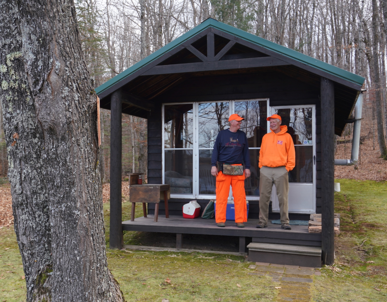 Two men in blaze orange stand on the porch of a small hunting cabin.
