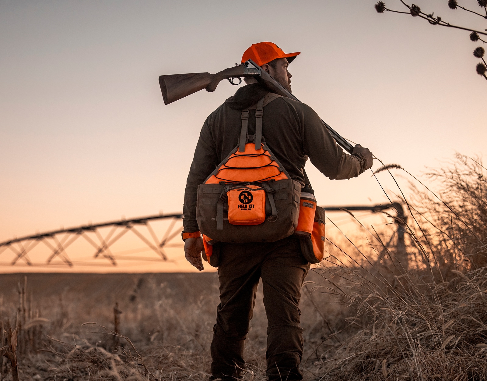 An upland hunter in blaze orange stands silhouetted against a sunset.