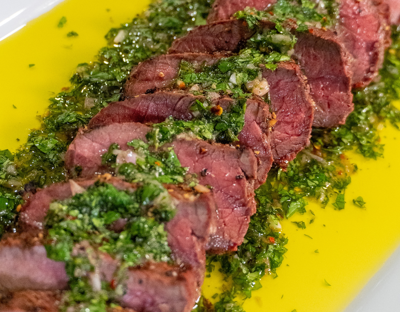 Elk backstraps grilled with a chimichuri sauce plated on a white platter.