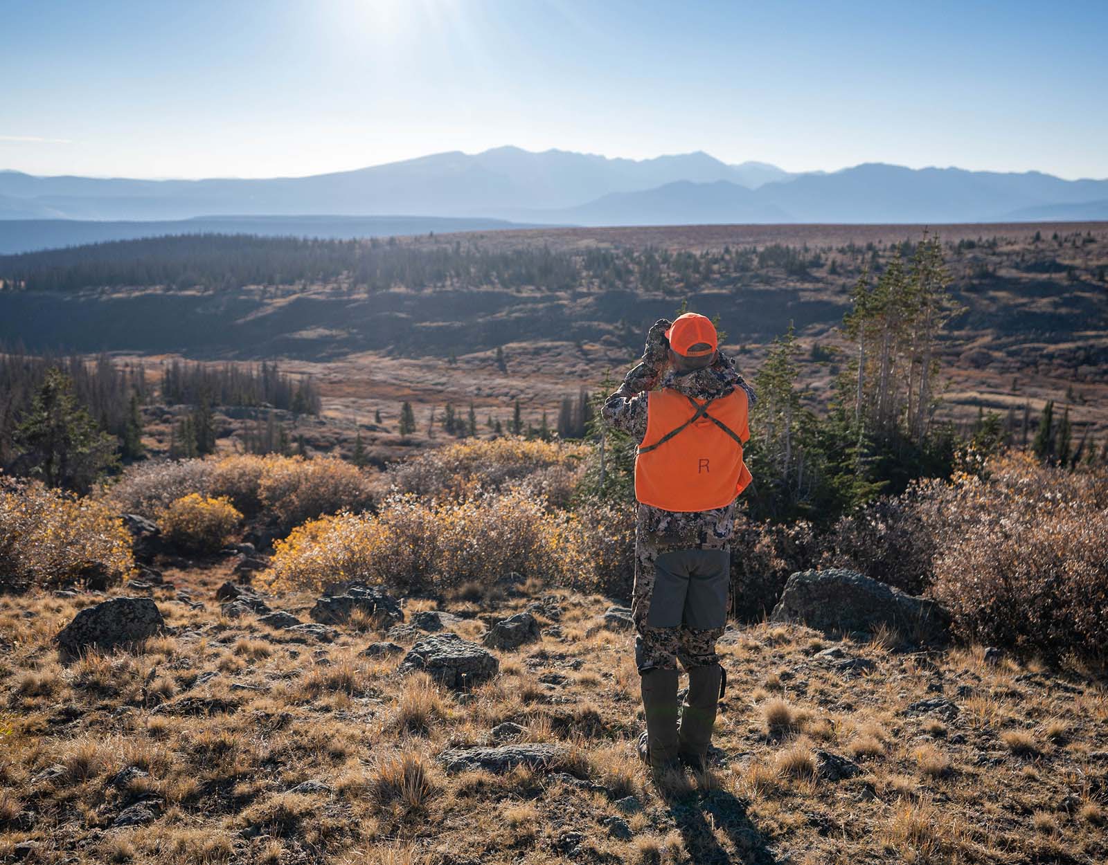 A hunter in blaze orange uses their binoculars to search sagebrush country.